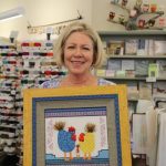 woman holding finished, framed canvas inside store with blue and yellow chickens with white baby chick with 3D embellishments and purple, green, and blue southwestern patterned border
